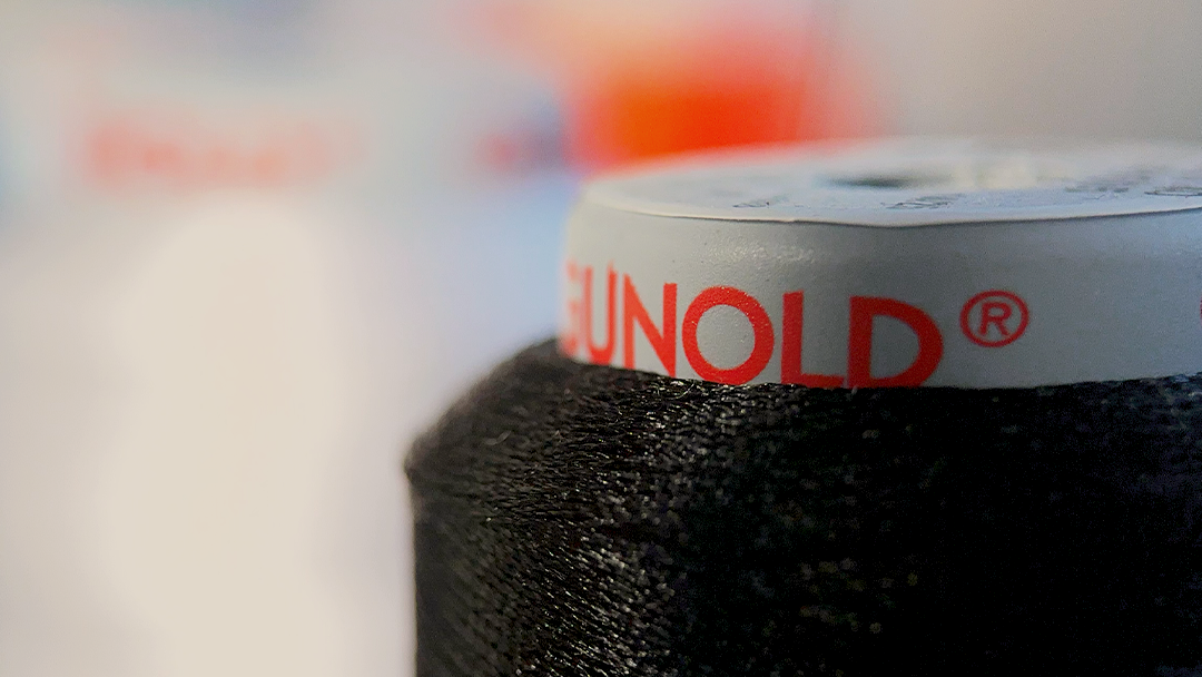 Nahaufnahme einer Spule mit schwarzem, glänzendem Stickgarn der Marke Gunold. Der Markenname ist in roten Großbuchstaben auf dem weißen oberen Rand der Rolle sichtbar. Der Hintergrund ist weich gezeichnet (Bokeh-Effekt).