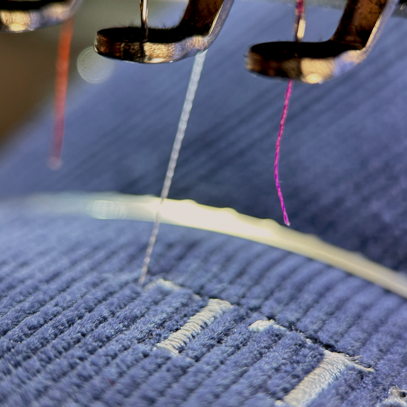 Nahaufnahme der MyStitch-Bestickung: Eine industrielle Mehrnadel-Stickmaschine beim Präzisions-Sticken eines dunkelblauen gerippten Stoffes mit weißem Faden. Metall-Nadelhalter und mehrere Fäden (weiß, magenta) sind im Fokus, während ein gefülltes Designelement entsteht. Textilveredelung aus Langenthal.