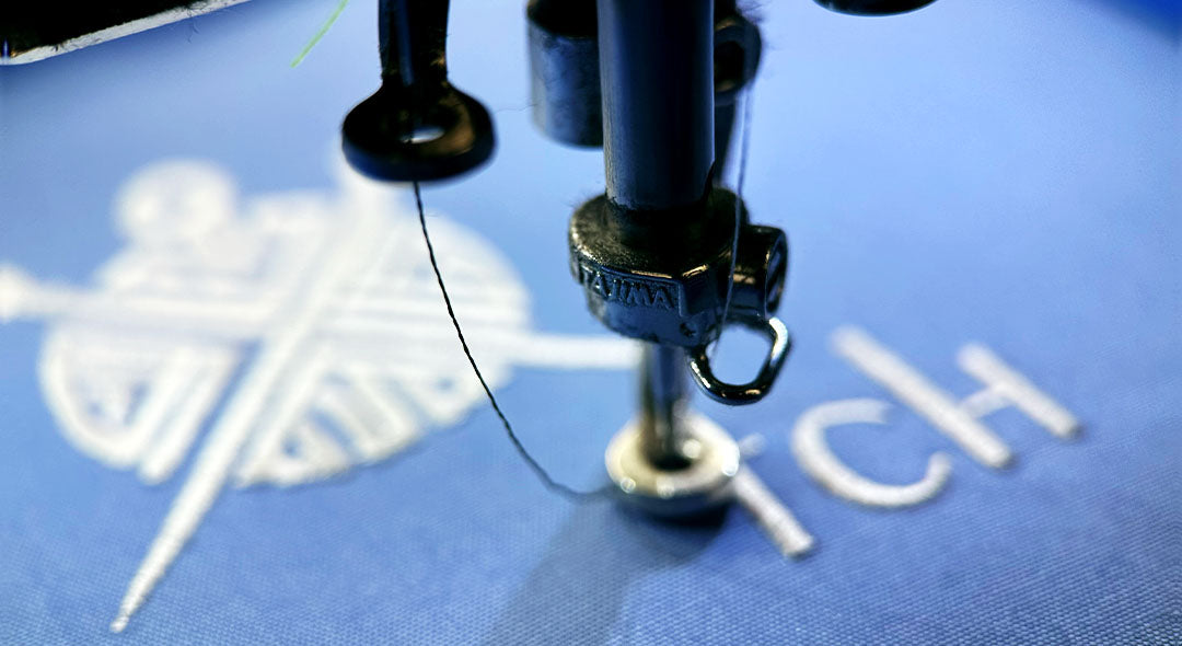 Nahaufnahme einer Tajima-Stickmaschine, die ein weißes Logo und den Schriftzug MyStitch auf blauen Stoff stickt.