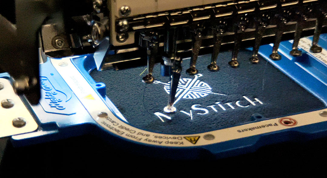 Hochwertige Textilveredelung: Eine professionelle Stickmaschine bestickt ein dunkelblaues Textil mit dem weissen MyStitch-Logo.