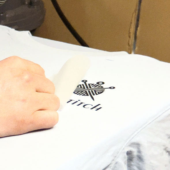 Nahaufnahme einer Hand, die eine klare Schutzfolie von einem weißen T-Shirt abzieht. Dieser Schritt zeigt den Abschluss eines Wärmeübertragungsprozesses (wie DTF), bei dem ein bedrucktes schwarzes Logo, bestehend aus einem geometrischen Nadel-und-Garn-Symbol und dem Text "titch", auf den Stoff übertragen wird.