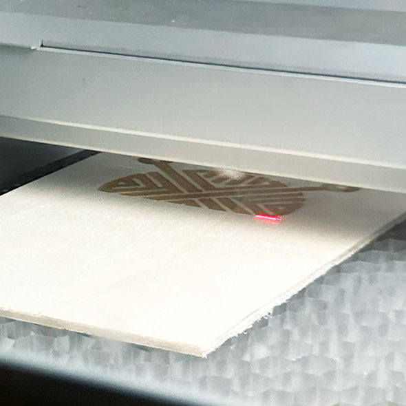 Eine Lasergravurmaschine in Langenthal graviert ein geometrisches Korbgeflecht-Muster in eine dünne Sperrholzplatte auf einem Wabenbett, wobei ein roter Laserpunkt sichtbar ist.