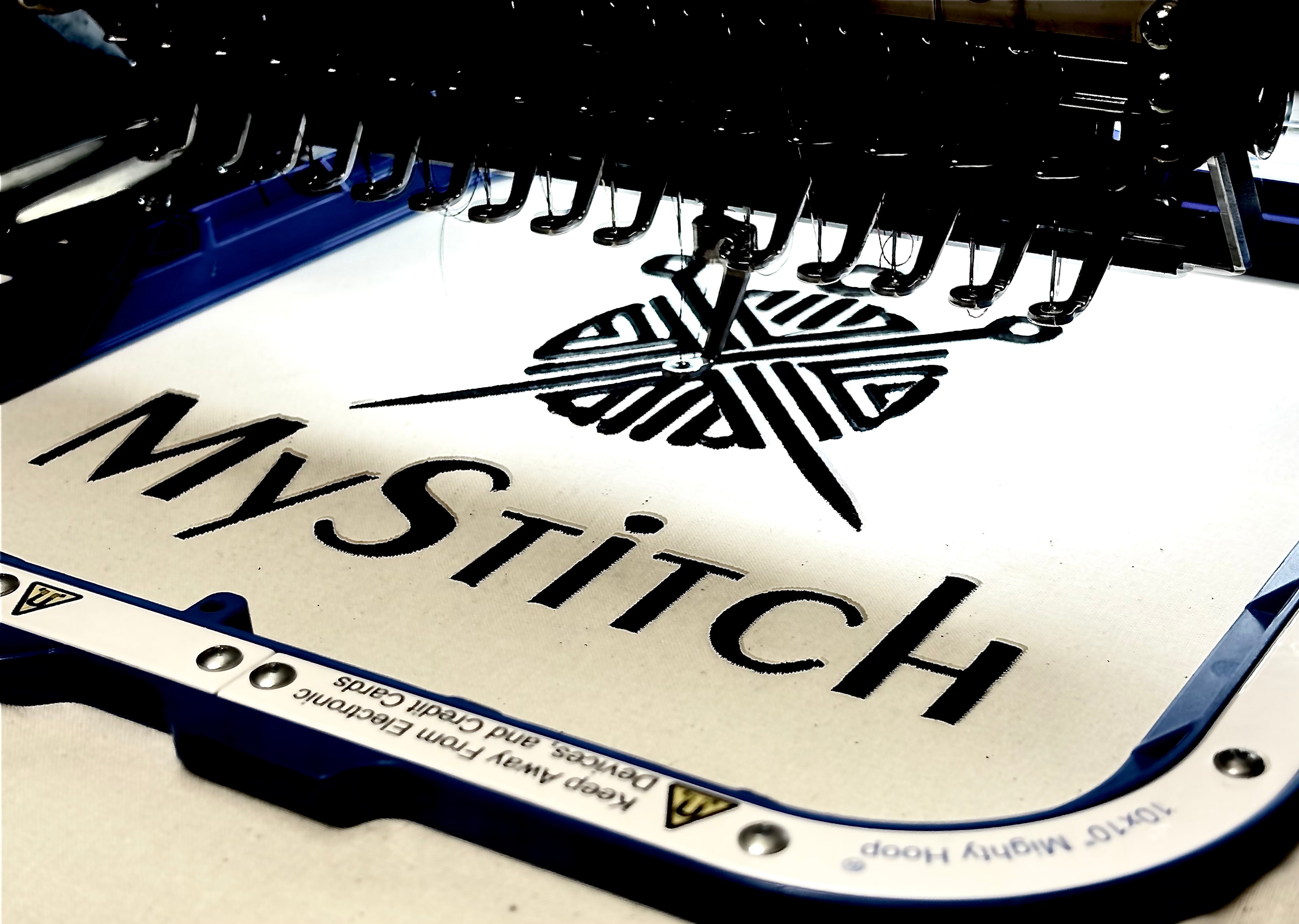 Nahaufnahme einer Stickmaschine im Einsatz. In einem blauen „Mighty Hoop“-Stickrahmen ist weißer Stoff eingespannt. Die Nadel stickt gerade ein schwarzes Logo, das aus einem Kreis mit Linien und einer Nadel besteht, über dem Schriftzug „MyStitch“. Im oberen Bereich sind die zahlreichen Garnführungen und Nadeln der Maschine vor dunklem Hintergrund erkennbar.