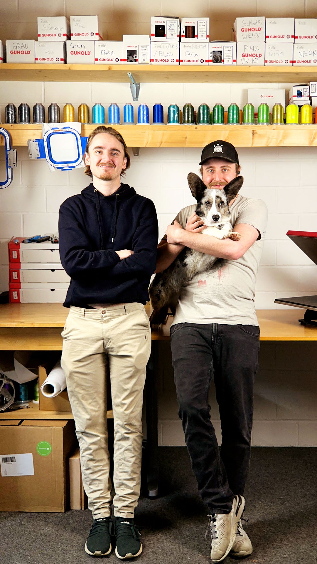 Ganzkörper-Fotografie von Nick und Alex Schüpbach aus Langenthal, dem MyStitch.ch Team, in ihrer Stickwerkstatt. Die beiden Männer stehen frontal vor Holzregalen, die mit farbenfrohen Stickgarnrollen, Boxen der Marke "GUNOLD" und Stickrahmen gefüllt sind. Nick Schüpbach (links) trägt einen schwarzen Hoodie und beige Chinos, während Alex Schüpbach (rechts) eine Kappe und schwarze Jeans trägt und lächelnd einen Corgi-Mischling im Arm hält. Die Werkstatt ist hell, ordentlich und professionell.